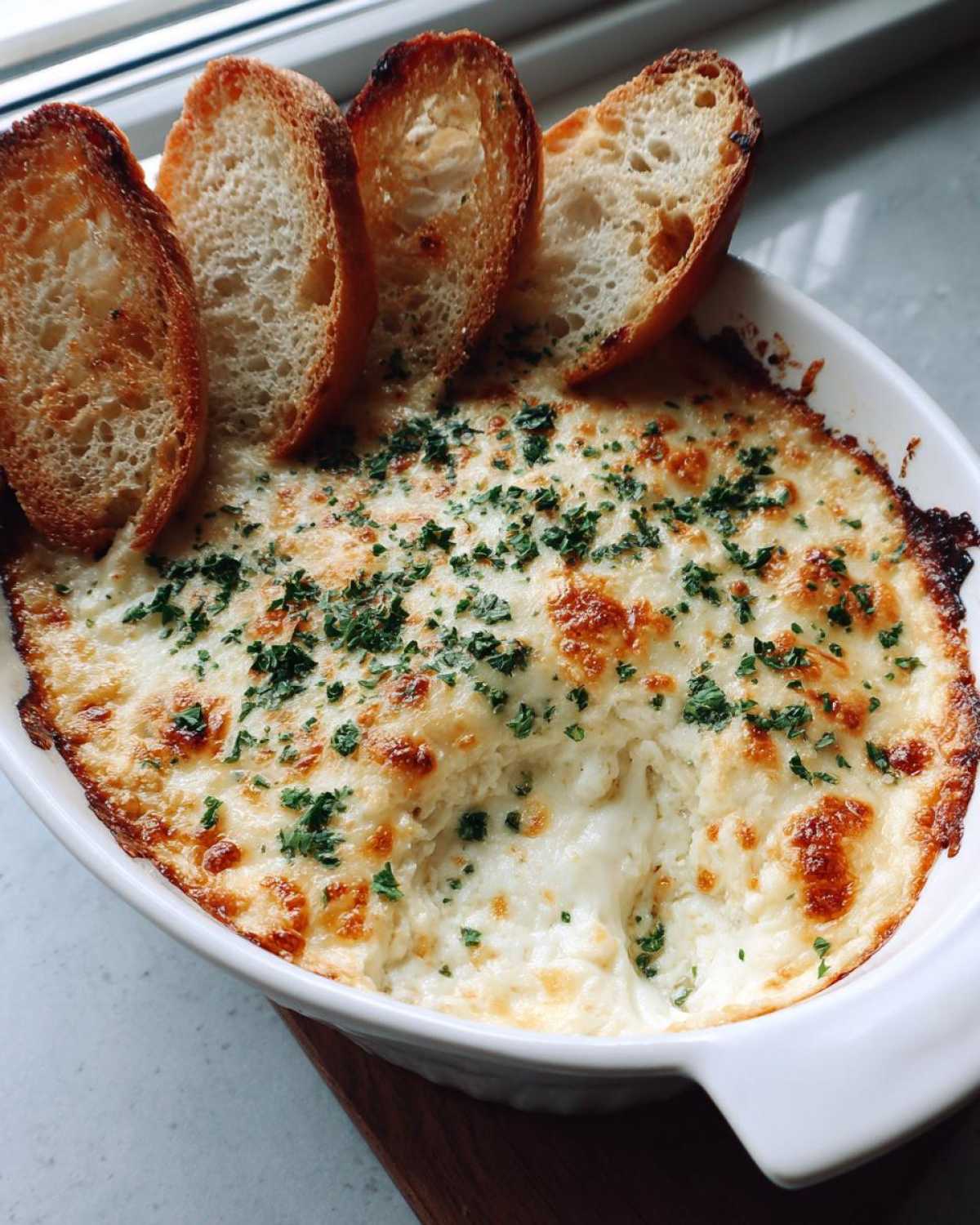 A bubbling, cheesy bread dip served hot in a white dish with toasted baguette slices for dipping.