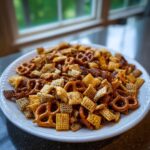 A large white platter piled high with homemade chex party mix featuring pretzels and seasoned cereal squares.