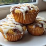 A stack of four freshly baked cinnamon roll muffins topped with thick white icing, sitting on a white plate.