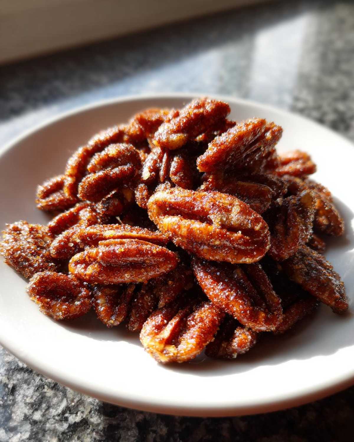 A small white bowl filled with glistening, candied cinnamon sugar pecans, highlighted by strong sunlight.