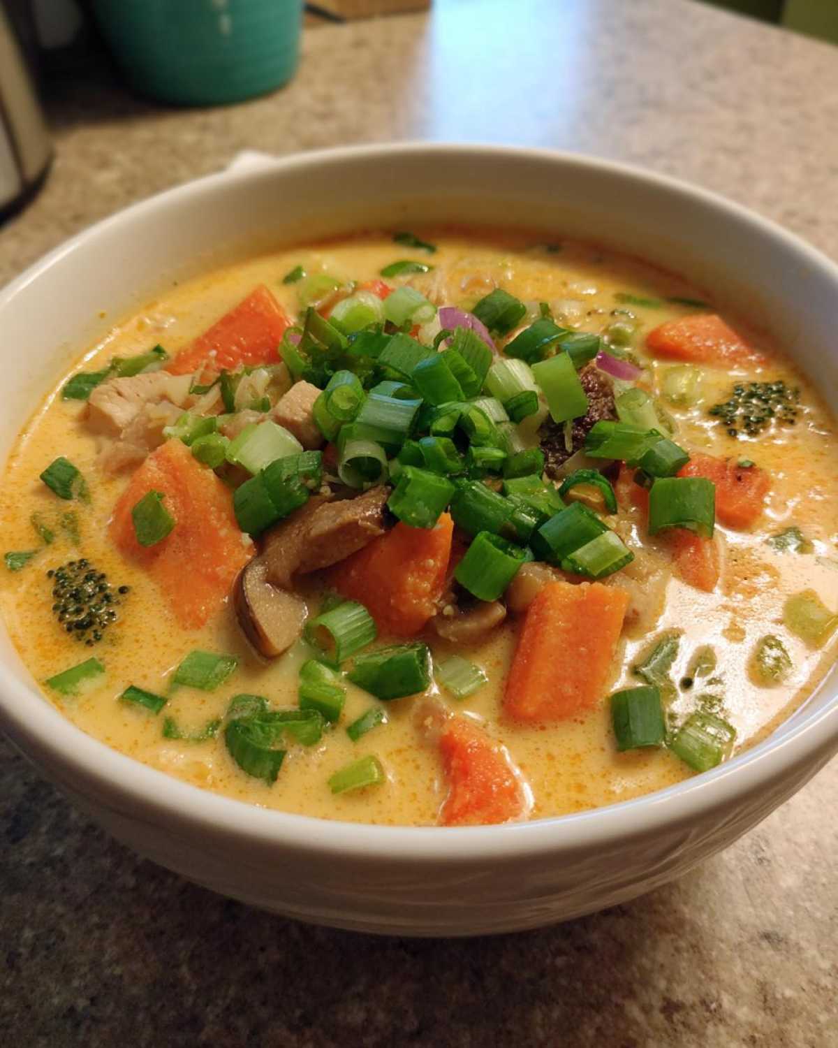 Close-up of a white bowl filled with creamy coconut curry soup, featuring chunks of carrot, mushrooms, and topped with fresh green onions.