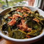 Close-up of a white bowl filled with savory, slow-cooked collard greens topped with pulled smoked meat.