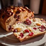 A slice of freshly baked cranberry walnut bread resting on a white plate next to the main loaf.