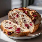 A loaf of freshly baked cranberry walnut bread, partially sliced, showing the soft interior studded with bright red cranberries and walnuts.