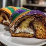 Close-up of a slice of cream cheese king cake showing swirled dough, filling, and purple, green, and gold sanding sugar.