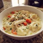 A close-up of a white bowl filled with creamy chicken and dumplings, topped with shredded chicken, carrots, and parsley.