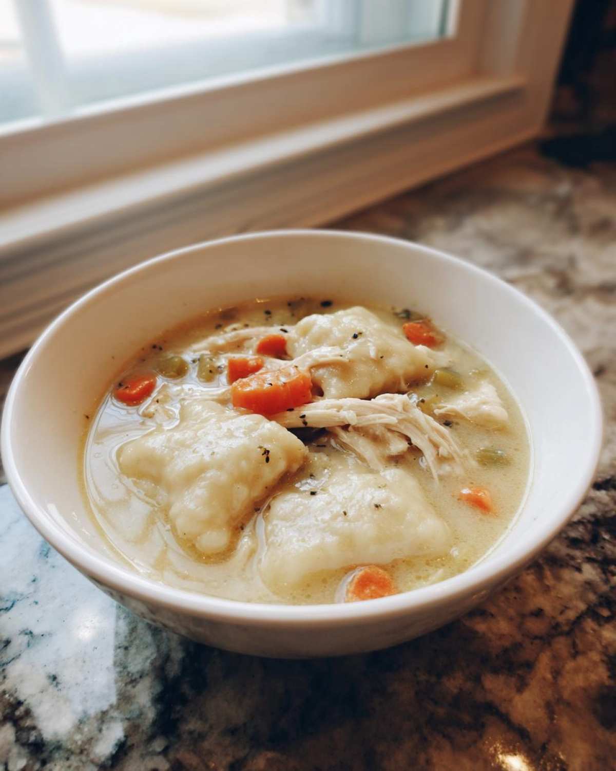 A white bowl filled with creamy chicken and dumplings, featuring shredded chicken, carrots, and herbs.