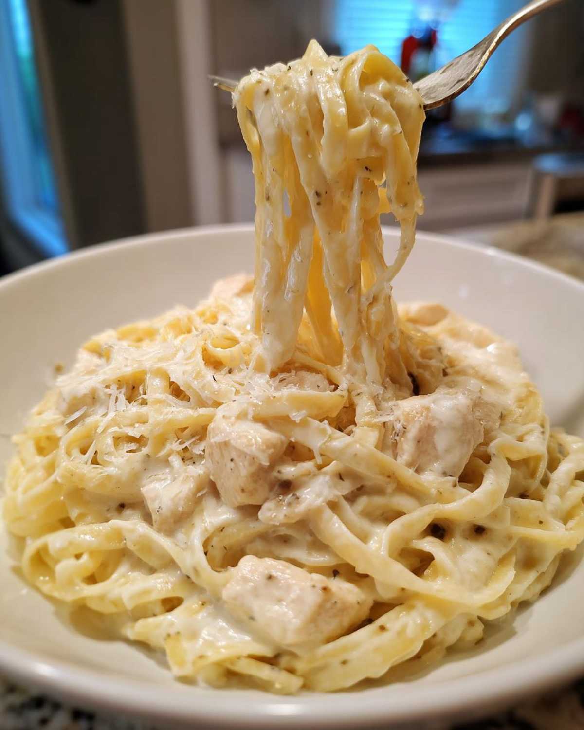 A fork lifting strands of creamy chicken fettuccine alfredo from a white bowl, showing chunks of chicken and grated Parmesan.