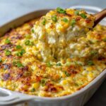 A spoonful of hot, cheesy, creamy corn dip being lifted from a baking dish, showing cheese pulls.