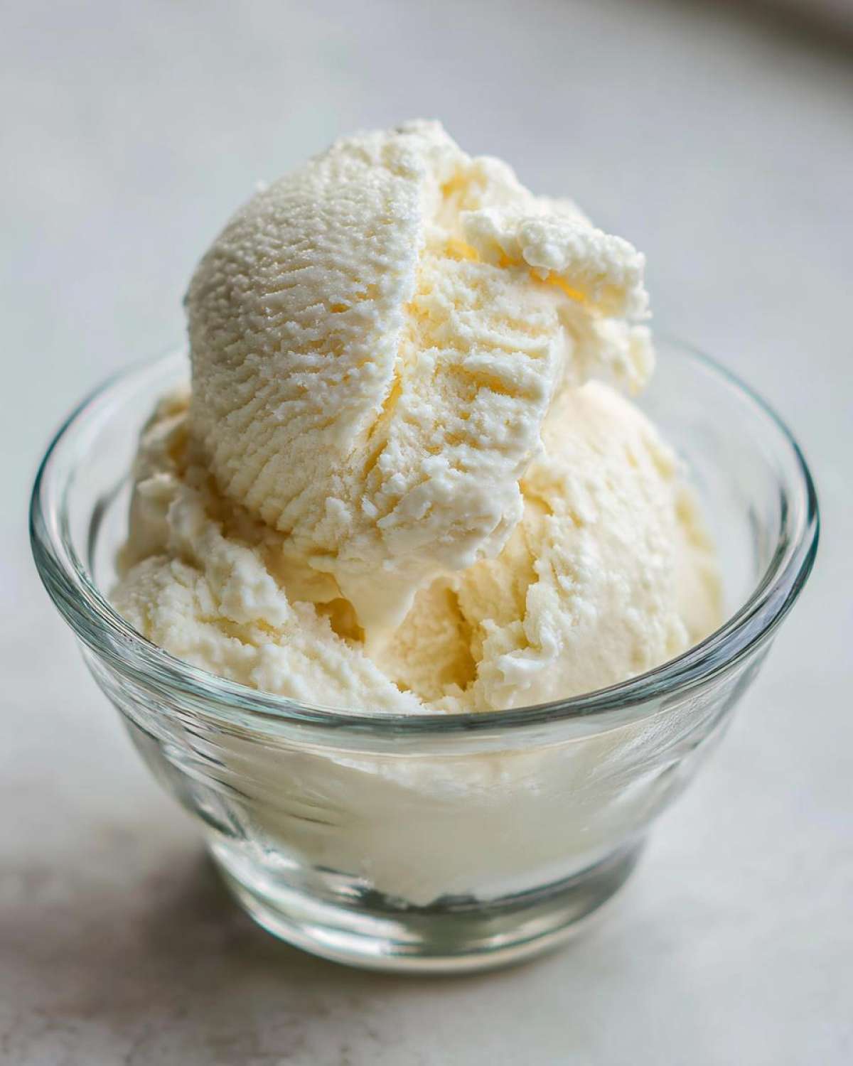 Two scoops of creamy, pale yellow cottage cheese ice cream served in a small clear glass bowl.