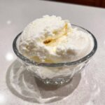 Two scoops of creamy, white cottage cheese ice cream served in a small, clear glass bowl.
