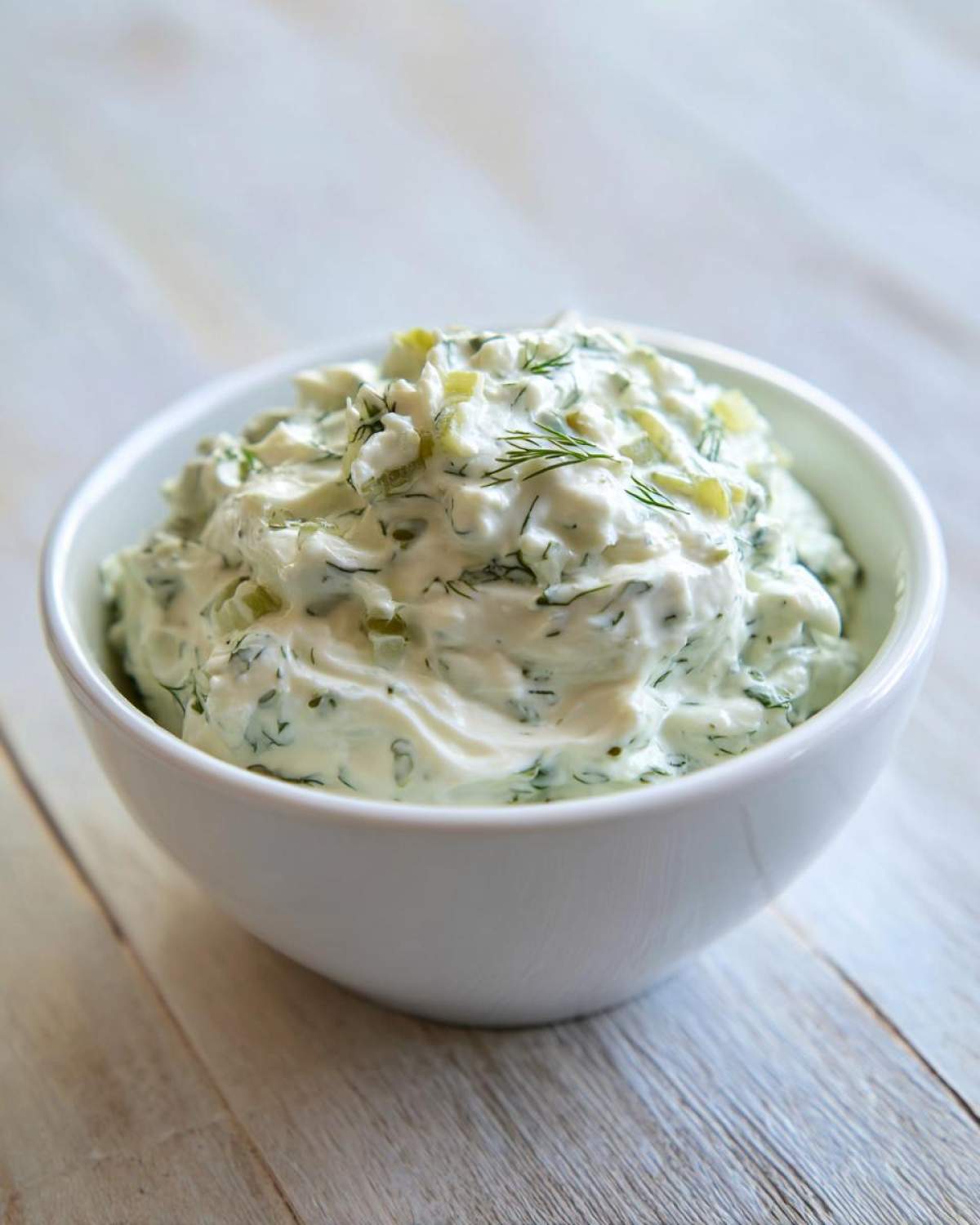 Close-up of creamy dill pickle dip, studded with chopped pickles and fresh dill, served in a white bowl.