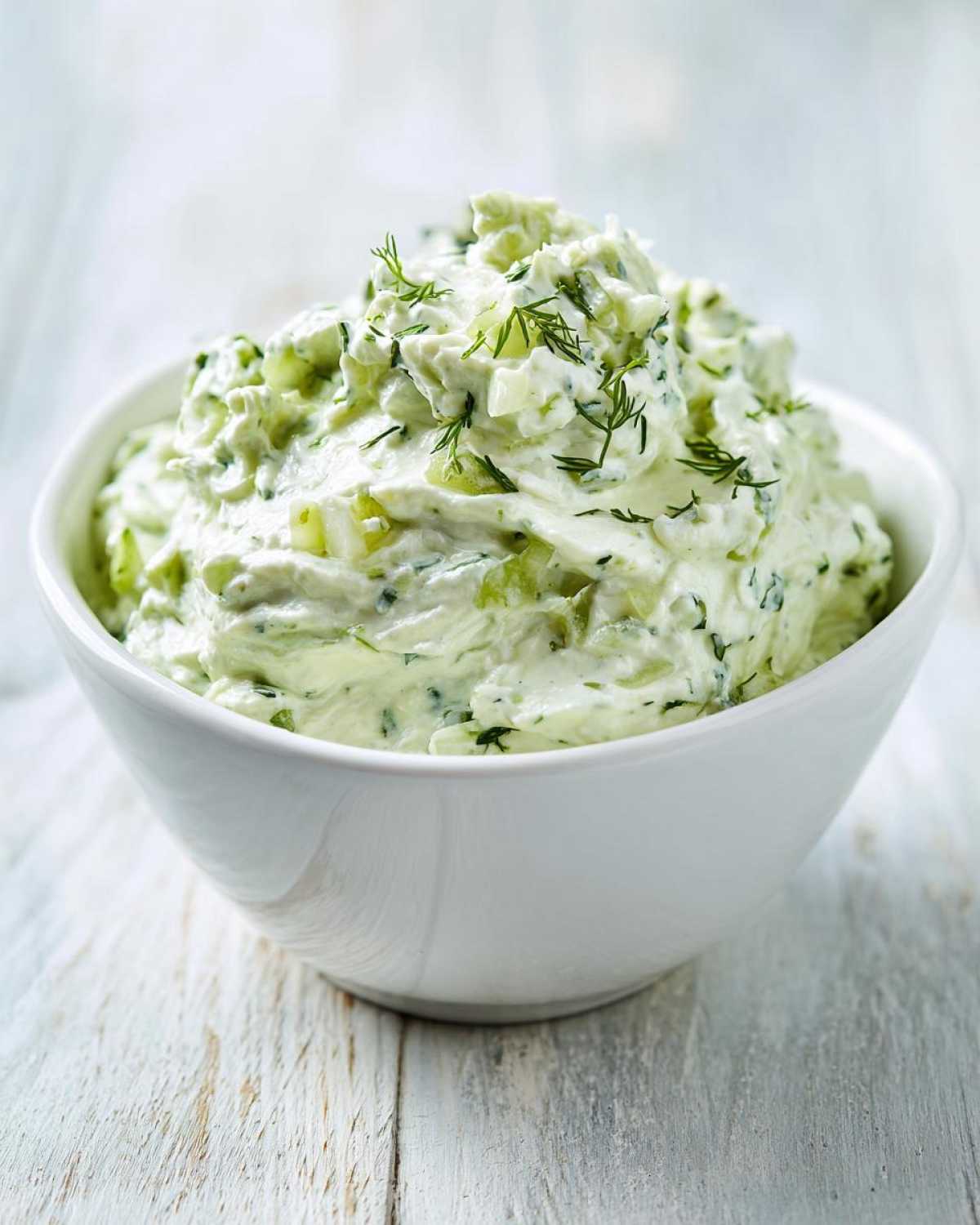 A close-up of creamy dill pickle dip, topped with fresh dill, served in a white bowl.