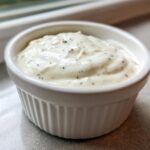 Close-up of thick, creamy garlic sauce speckled with herbs served in a small white fluted ramekin.