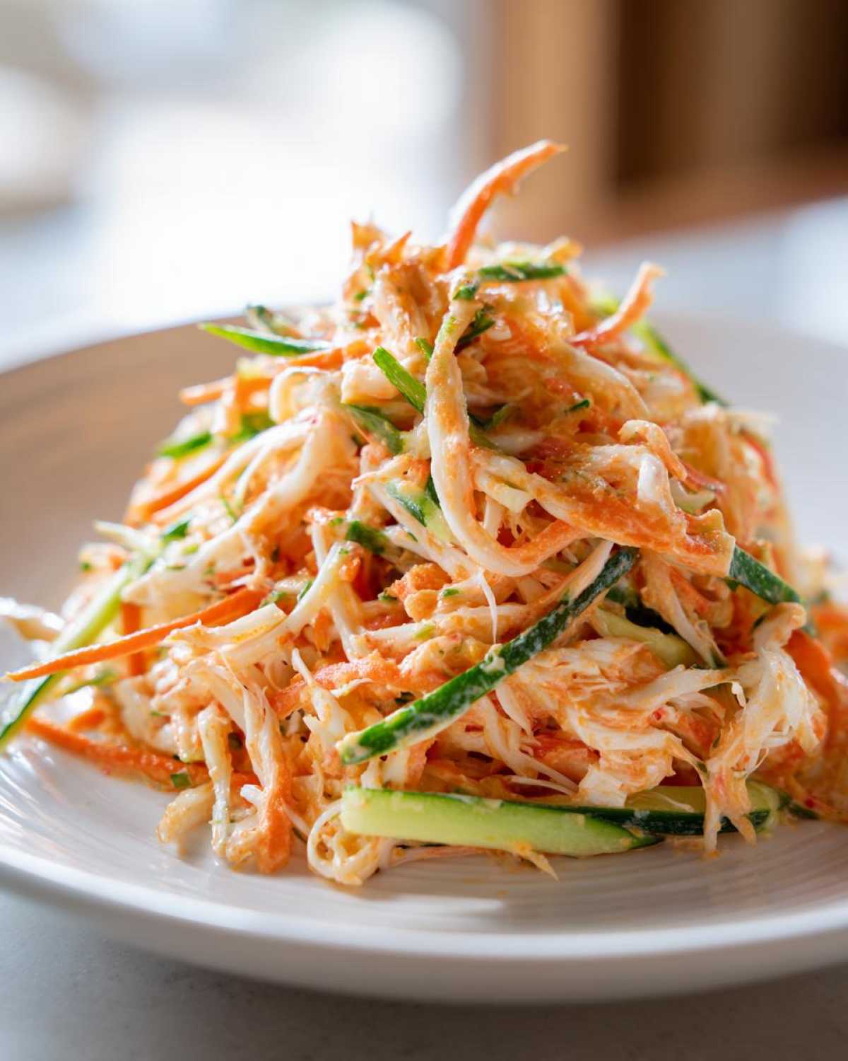 A mound of creamy kani salad featuring shredded imitation crab, julienned cucumber, and a light orange dressing on a white plate.