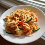 A close-up of a creamy kani salad featuring shredded imitation crab, julienned carrots, and cucumber slices on a white plate.
