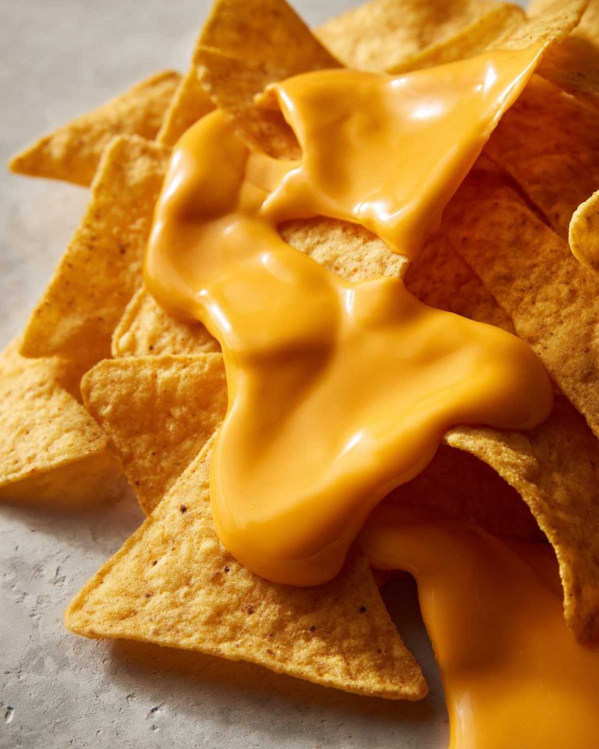 Close-up of creamy, bright orange nacho cheese being poured over a pile of golden tortilla chips.