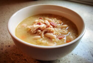 Close-up of a creamy bowl of navy bean soup topped with shredded chicken and ham pieces.