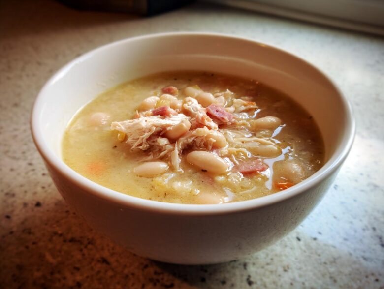 Close-up of a creamy bowl of navy bean soup topped with shredded chicken and ham pieces.