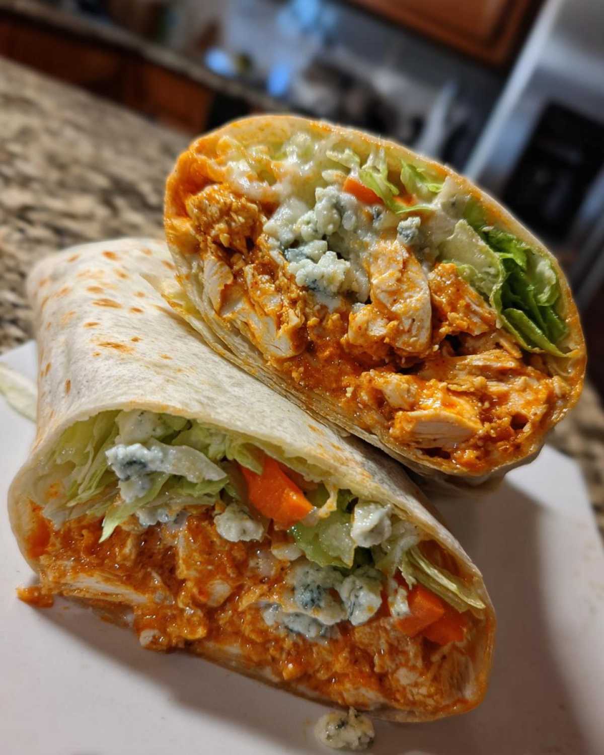 Close-up of a buffalo chicken wrap cut in half, showing spicy chicken, blue cheese crumbles, lettuce, and carrots.