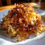 A close-up of a golden brown pile of crispy shredded fried potatoes glistening in the sunlight on a white plate.