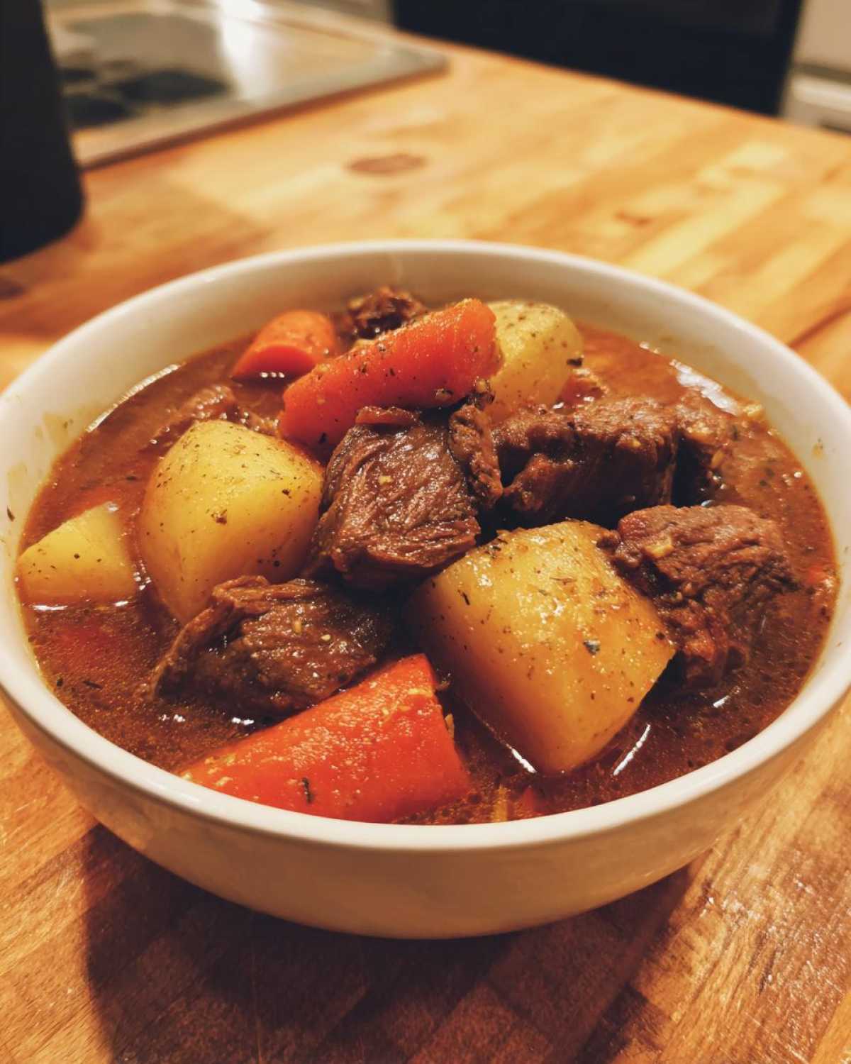 A close-up of a white bowl filled with rich crockpot beef stew, featuring tender beef chunks, potatoes, and carrots.