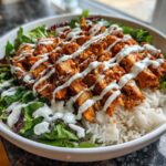 A close-up of a white bowl filled with white rice, mixed greens, and topped with sliced, spicy harissa chicken drizzled with white sauce.