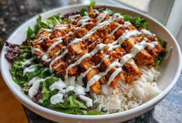 A close-up of a white bowl filled with white rice, mixed greens, and topped with sliced, spicy harissa chicken drizzled with white sauce.
