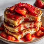 Close-up of a stack of three fluffy strawberry pancakes dripping with strawberry sauce and fresh slices.