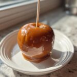 A single, glossy caramel apple on a stick, resting on a white plate, perfectly coated in rich caramel.