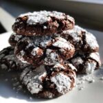 A stack of rich, dark chocolate crinkle cookies heavily dusted with white powdered sugar, sitting on a white plate.