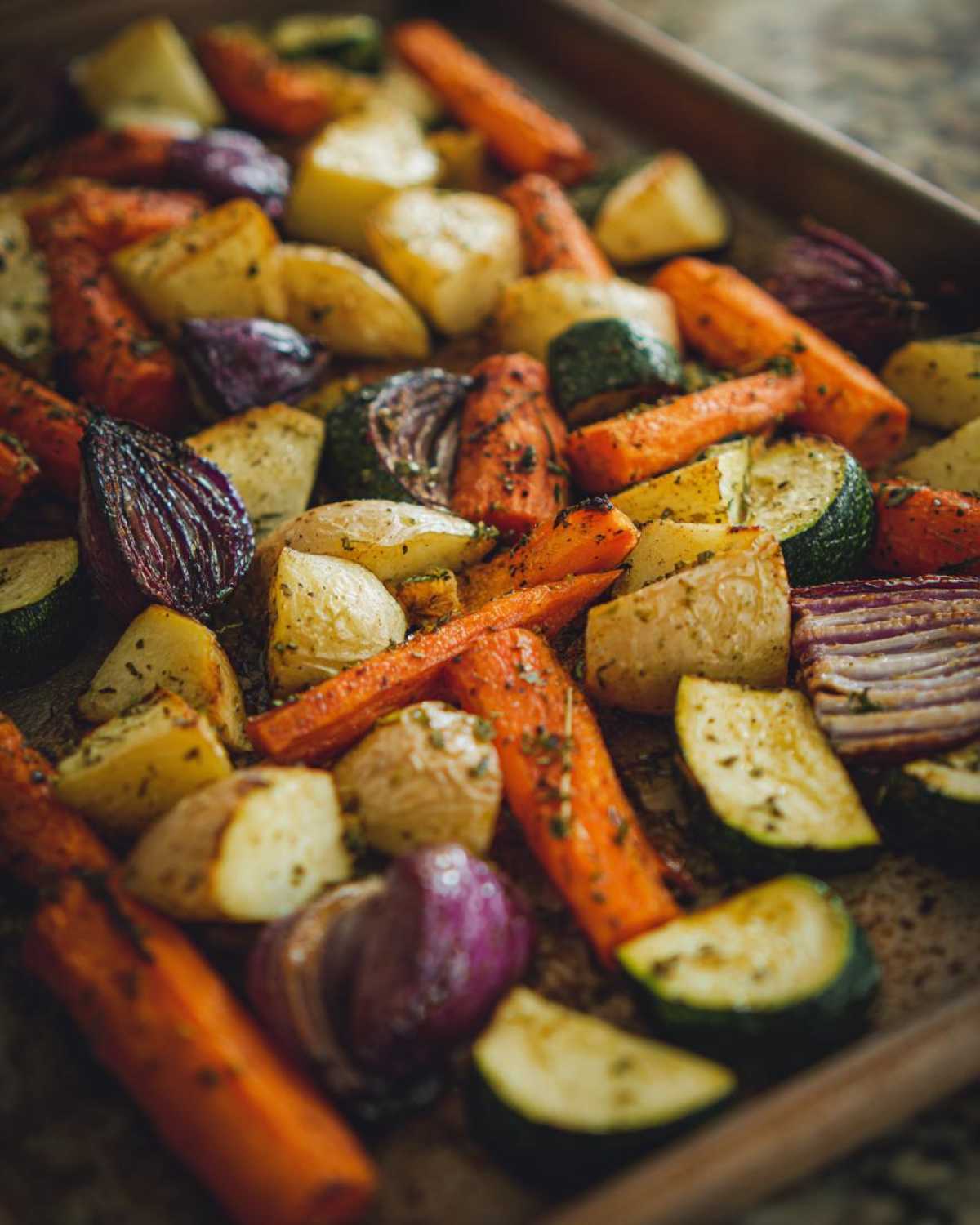 Close-up of colorful Garlic Herb Roasted Veggies including carrots, potatoes, zucchini, and red onion on a baking sheet.