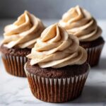 Three rich, dark chocolate gingerbread cupcakes topped with swirls of light brown cream cheese frosting dusted with spice.