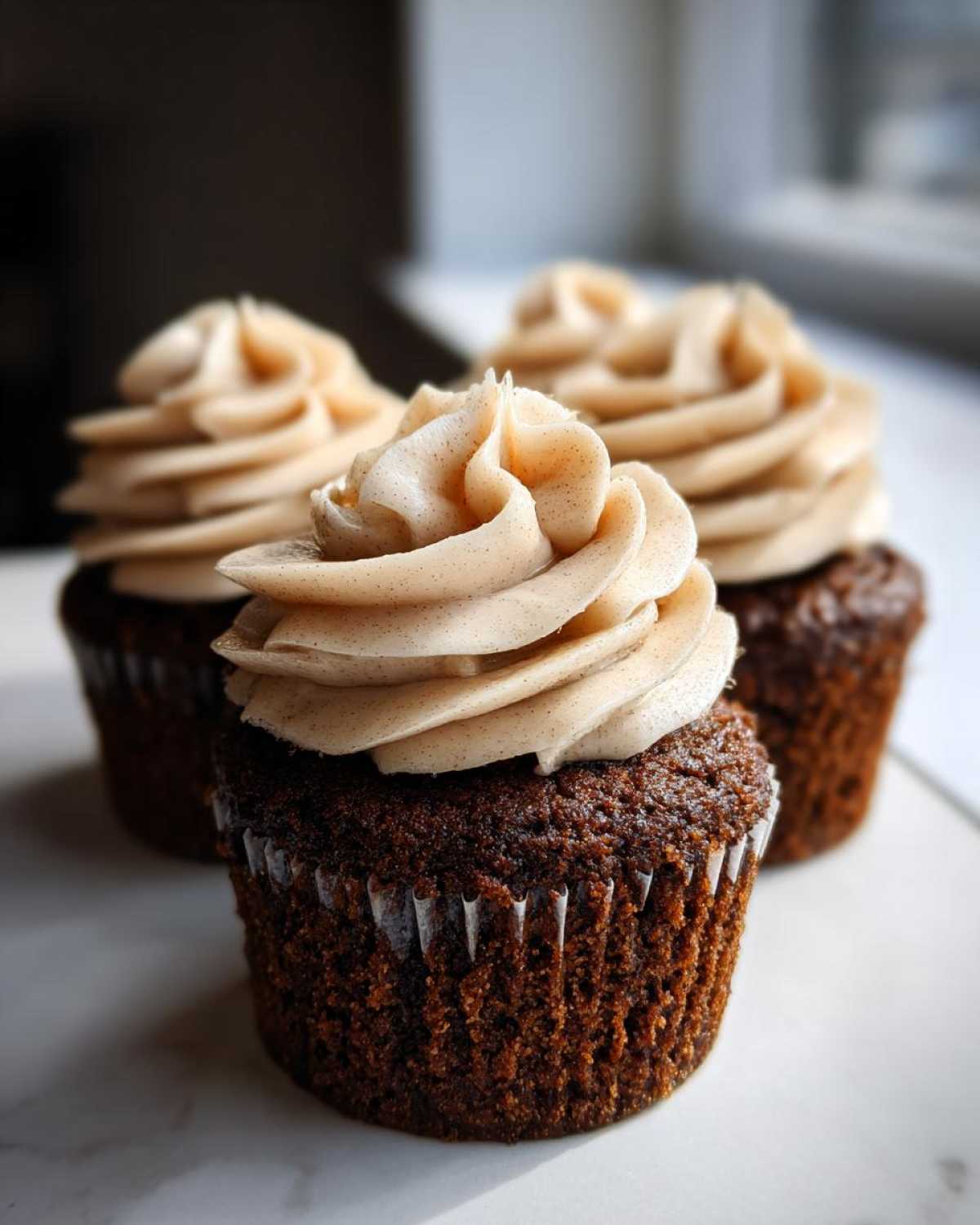 Three rich, dark gingerbread cupcakes topped with swirls of light brown, spiced cream cheese frosting.