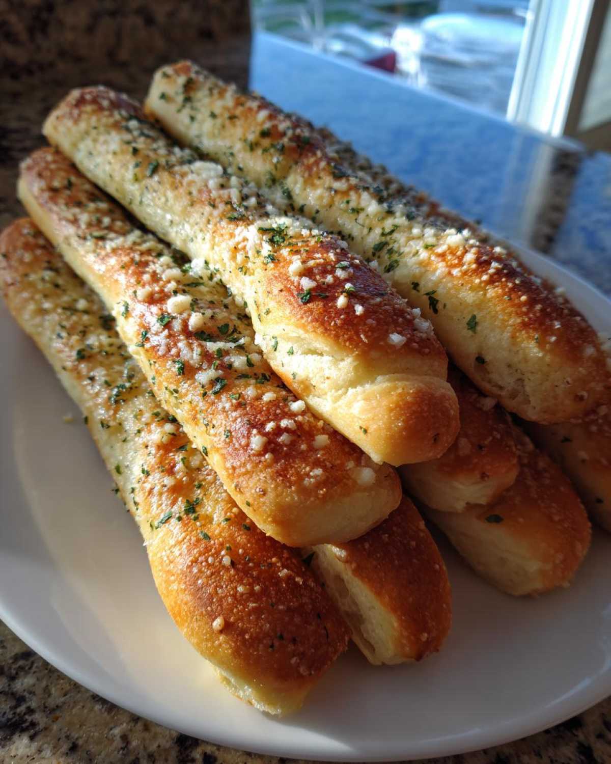 A pile of freshly baked, golden brown breadsticks topped with garlic, parmesan cheese, and parsley, served on a white plate.