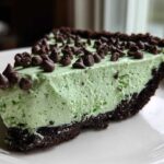 A close-up slice of bright green grasshopper pie with a dark chocolate crust, topped with mini chocolate chips.
