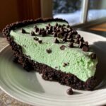 A delicious slice of grasshopper pie featuring a thick, light green mint filling and a dark chocolate crust, topped with mini chocolate chips.