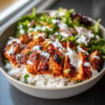 A close-up of a bowl featuring sliced, spiced harissa chicken served over white rice, drizzled with a white sauce, alongside a fresh green salad.