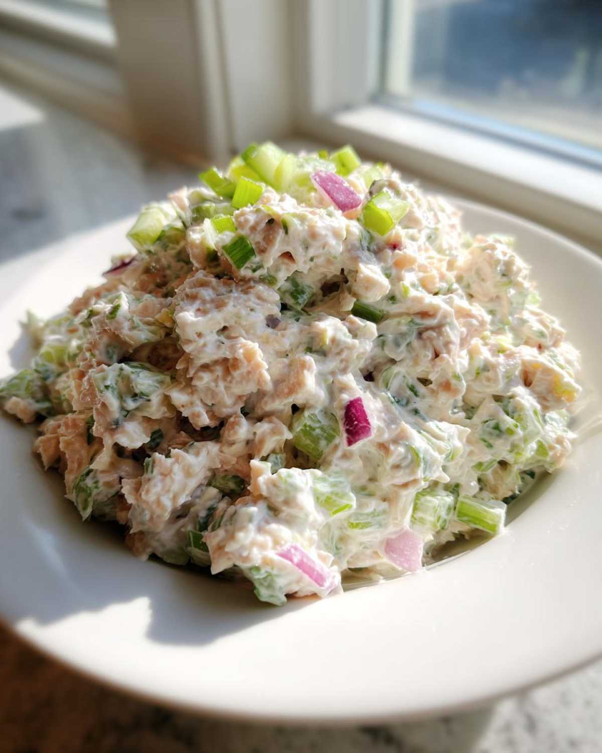 A mound of creamy healthy tuna salad mixed with chopped celery and red onion, served on a white plate.