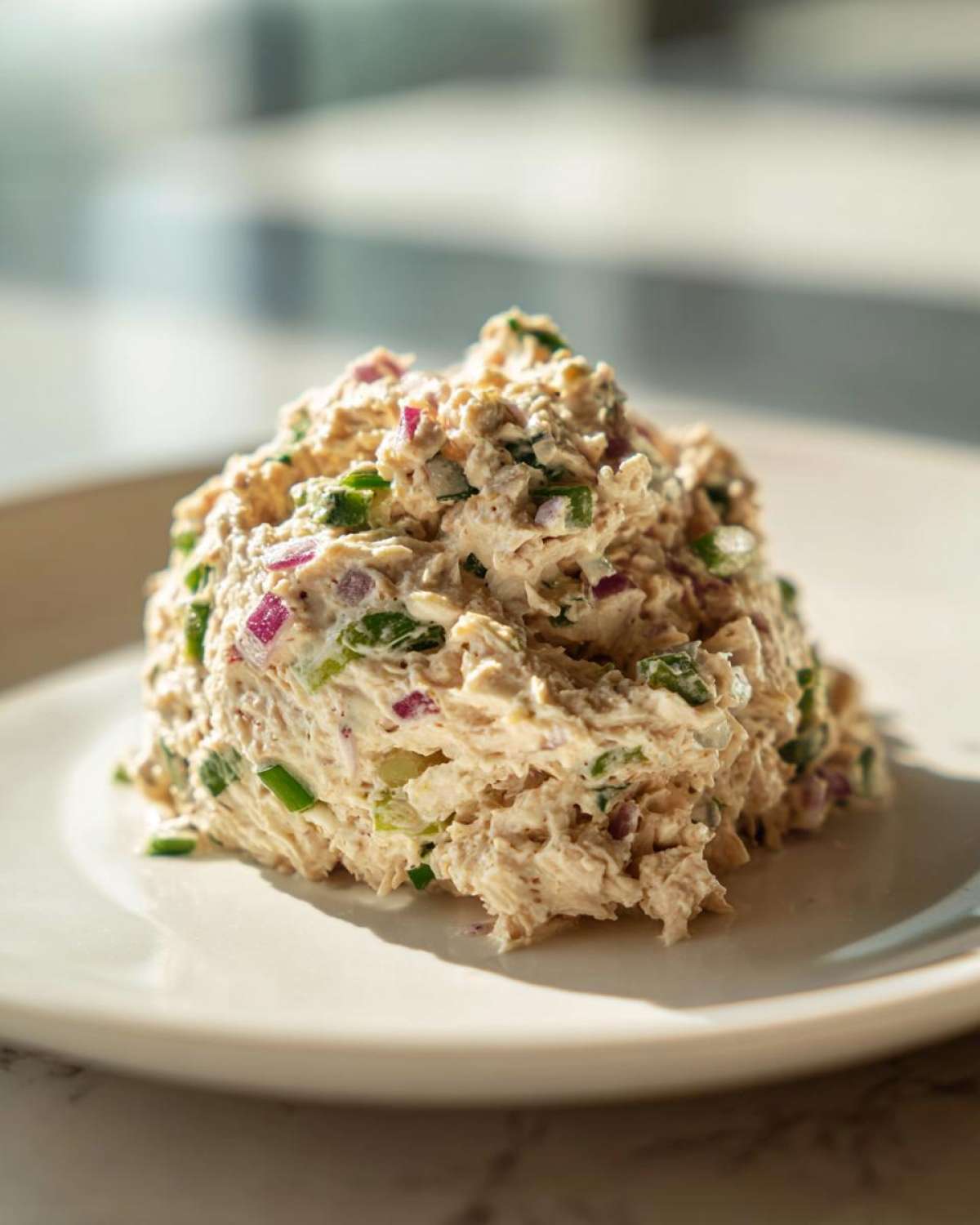A mound of creamy healthy tuna salad mixed with visible pieces of red onion and green onion on a white plate.