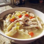 A close-up of a white bowl filled with steaming chicken and dumplings, featuring shredded chicken, carrots, and celery in broth.