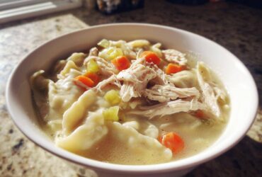 A close-up of a white bowl filled with steaming chicken and dumplings, featuring shredded chicken, carrots, and celery in broth.