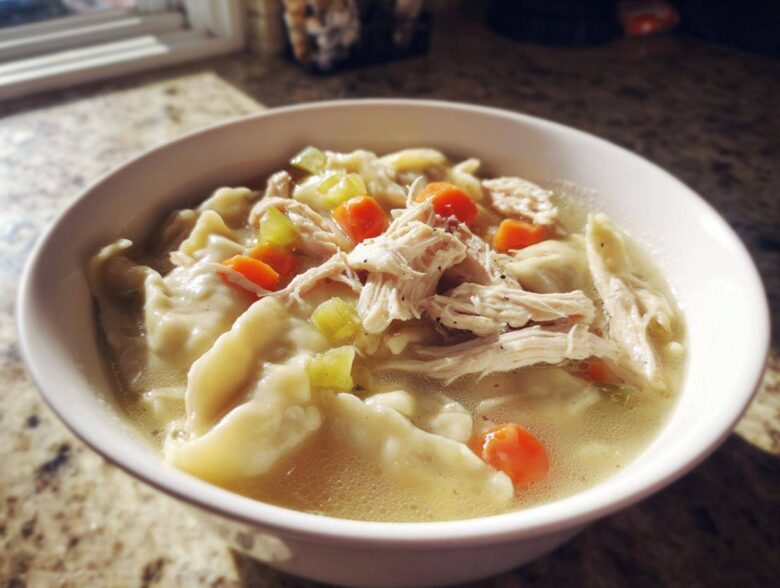 A close-up of a white bowl filled with steaming chicken and dumplings, featuring shredded chicken, carrots, and celery in broth.