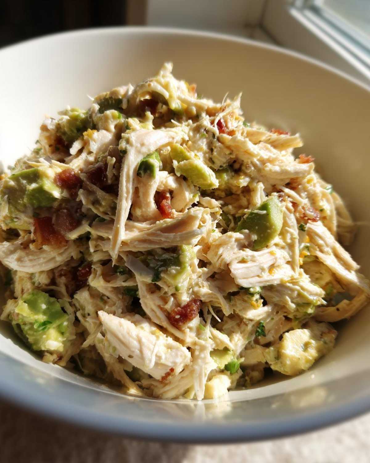 Close-up of creamy honey mustard chicken salad mixed with chunks of avocado and bits of bacon in a white bowl.