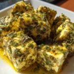 Close-up of several cubes of bright yellow marinated cheese heavily coated in green herbs and oil.