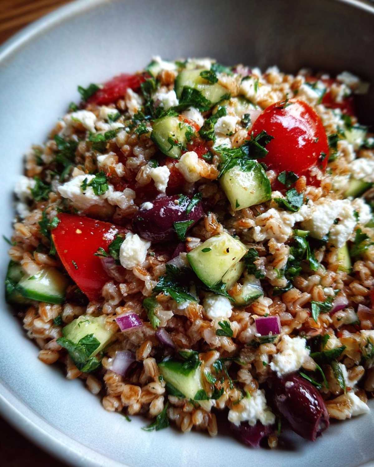 Close-up of a vibrant farro salad mixed with chopped cucumbers, cherry tomatoes, feta cheese, olives, and parsley.