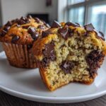 Close-up of two banana chocolate chip muffins, one broken open showing a moist interior packed with chocolate chunks.