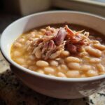 Close-up of a white bowl filled with rich navy bean soup topped with shredded ham.