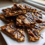 Close-up of stacked, square pieces of Pecan Pie Bark drizzled with dark chocolate on a white plate.