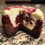 A close-up view showing the layers of a red velvet cheesecake, featuring a dark crust, rich red cake, and swirled cream cheese topping.